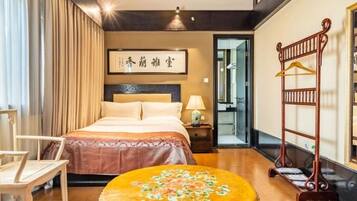 A bedroom with a bed, a nightstand, a chair, a round table with a floral tablecloth, and a wooden rack with hangers.