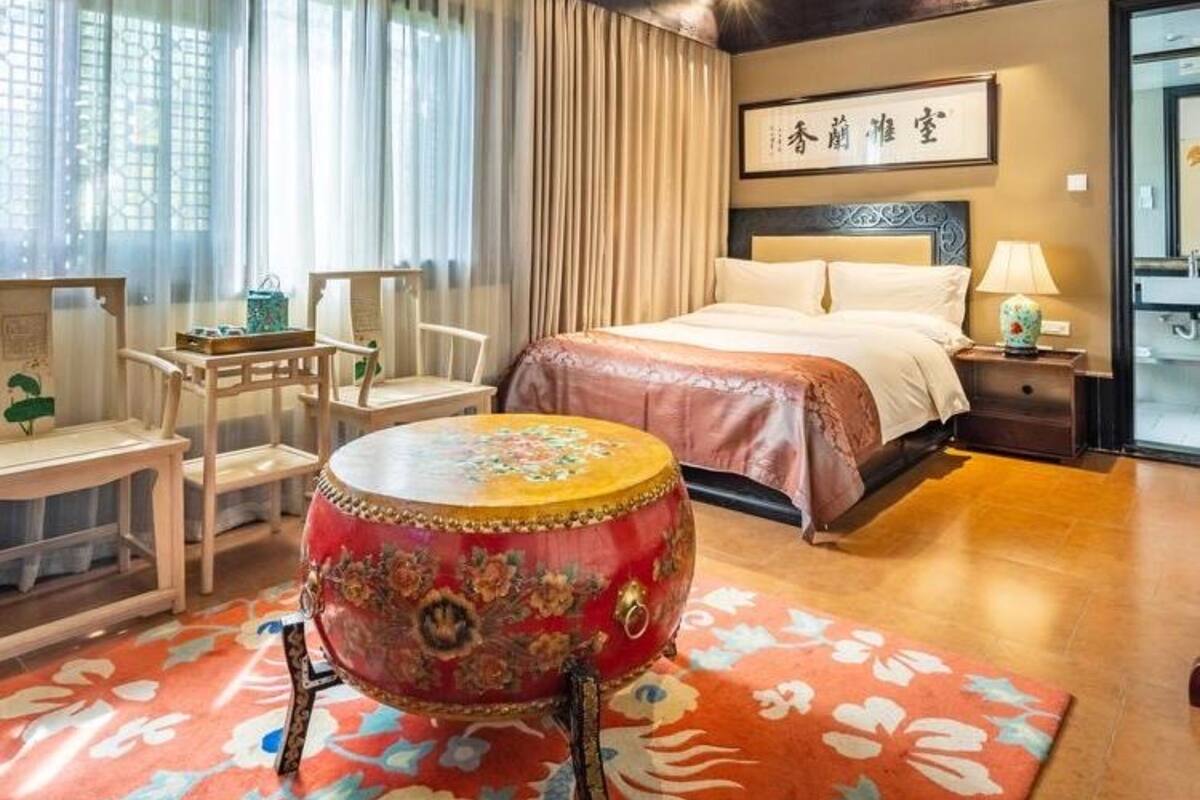 Une chambre à coucher de style traditionnel, comprenant un lit, un bureau, une chaise, un tambour décoratif et une grande fenêtre avec des rideaux.