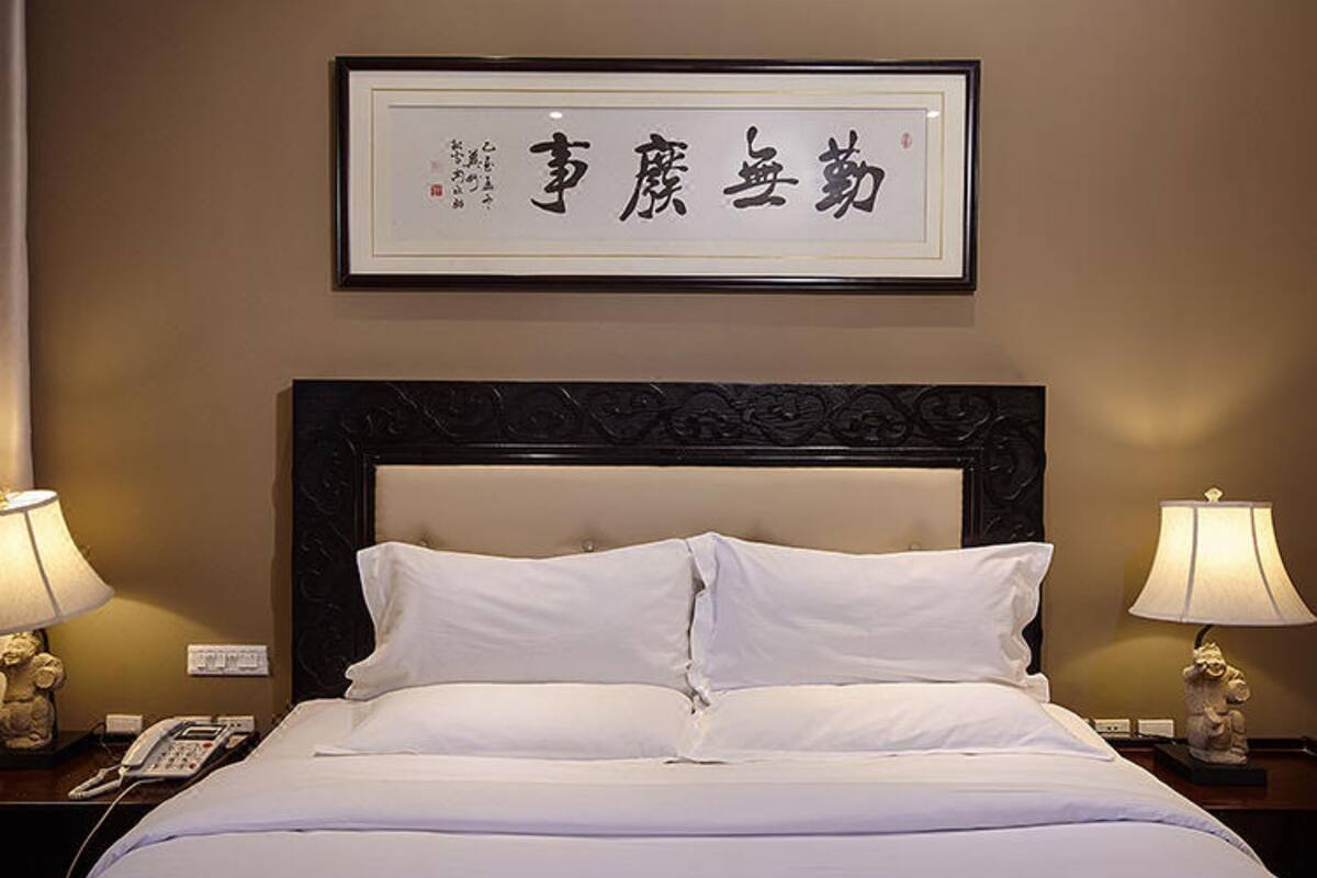 Une chambre d’hôtel avec un grand lit, deux lampes de chevet, une œuvre calligraphique encadrée accrochée au mur et un téléphone sur la table de chevet.
