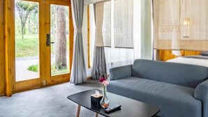 A modern room with a grey sofa, a small black table with a vase and remote, large windows with curtains, and a wooden ceiling.