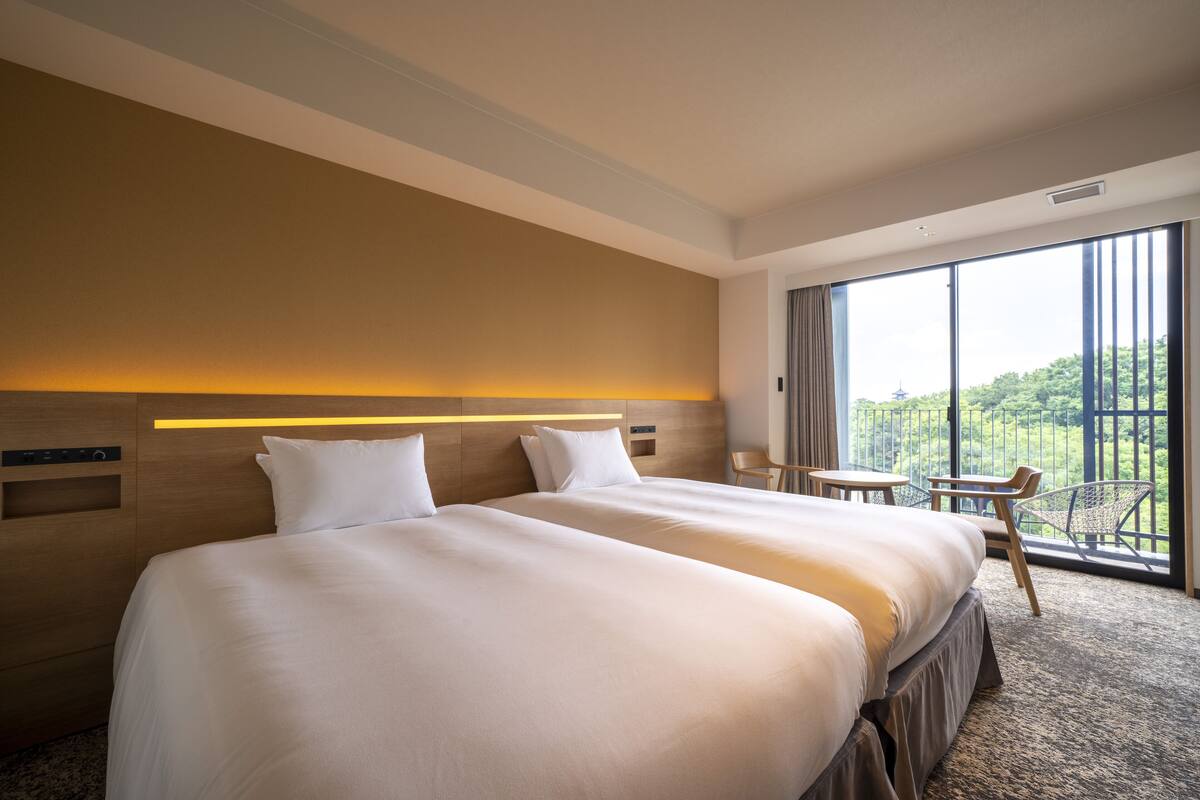 Habitación de hotel con dos camas, una mesita pequeña, una silla y un ventanal grande con vistas a zonas verdes.