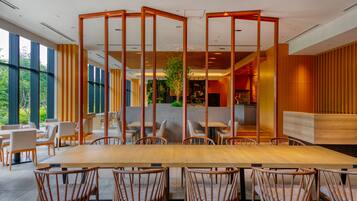 A modern restaurant with a large wooden table and chairs, a wooden partition, and large windows offering a view of greenery.