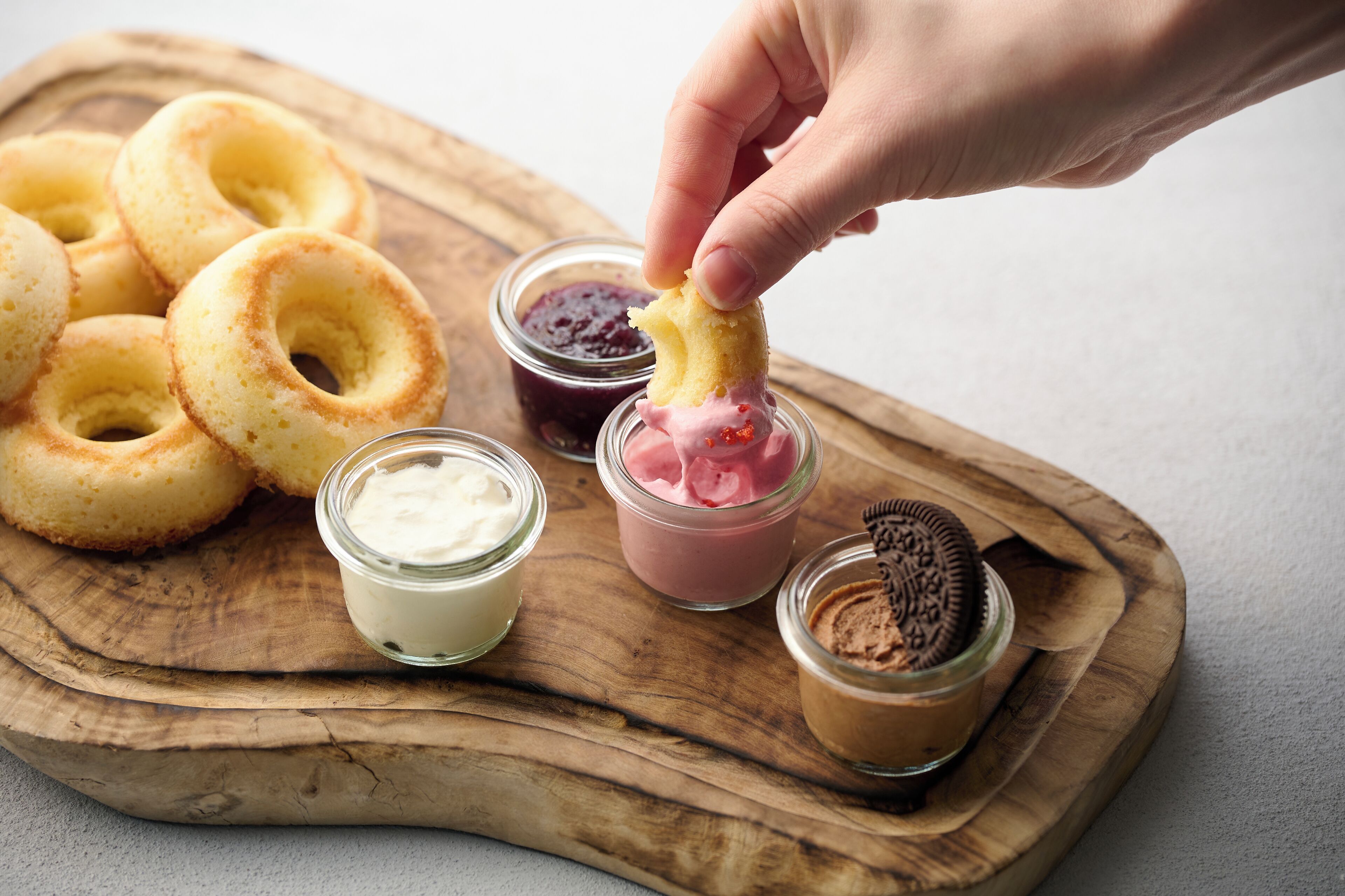 A wooden board with donuts, three small jars of different colored dips, and a hand dipping a donut into one of the jars.