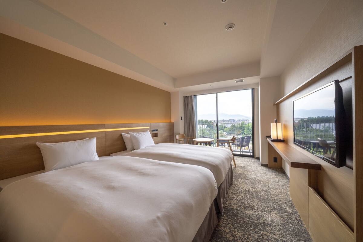 Una habitación de hotel con dos camas, televisión, escritorio y ventana grande con vista a la ciudad.