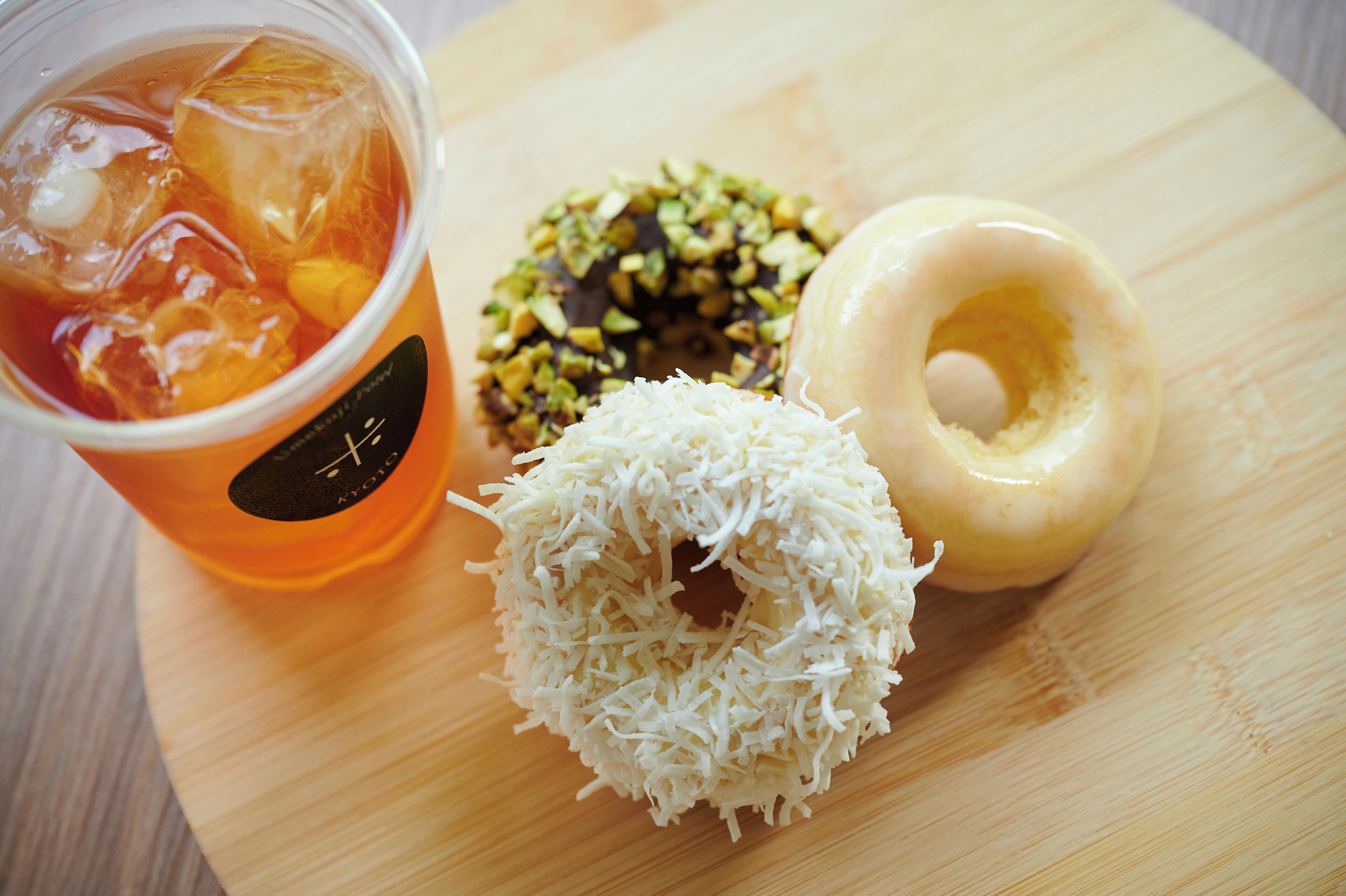 Een houten tafel met een drankje in een glas, een donut met witte glazuur en een chocoladedonut met groene toppings.