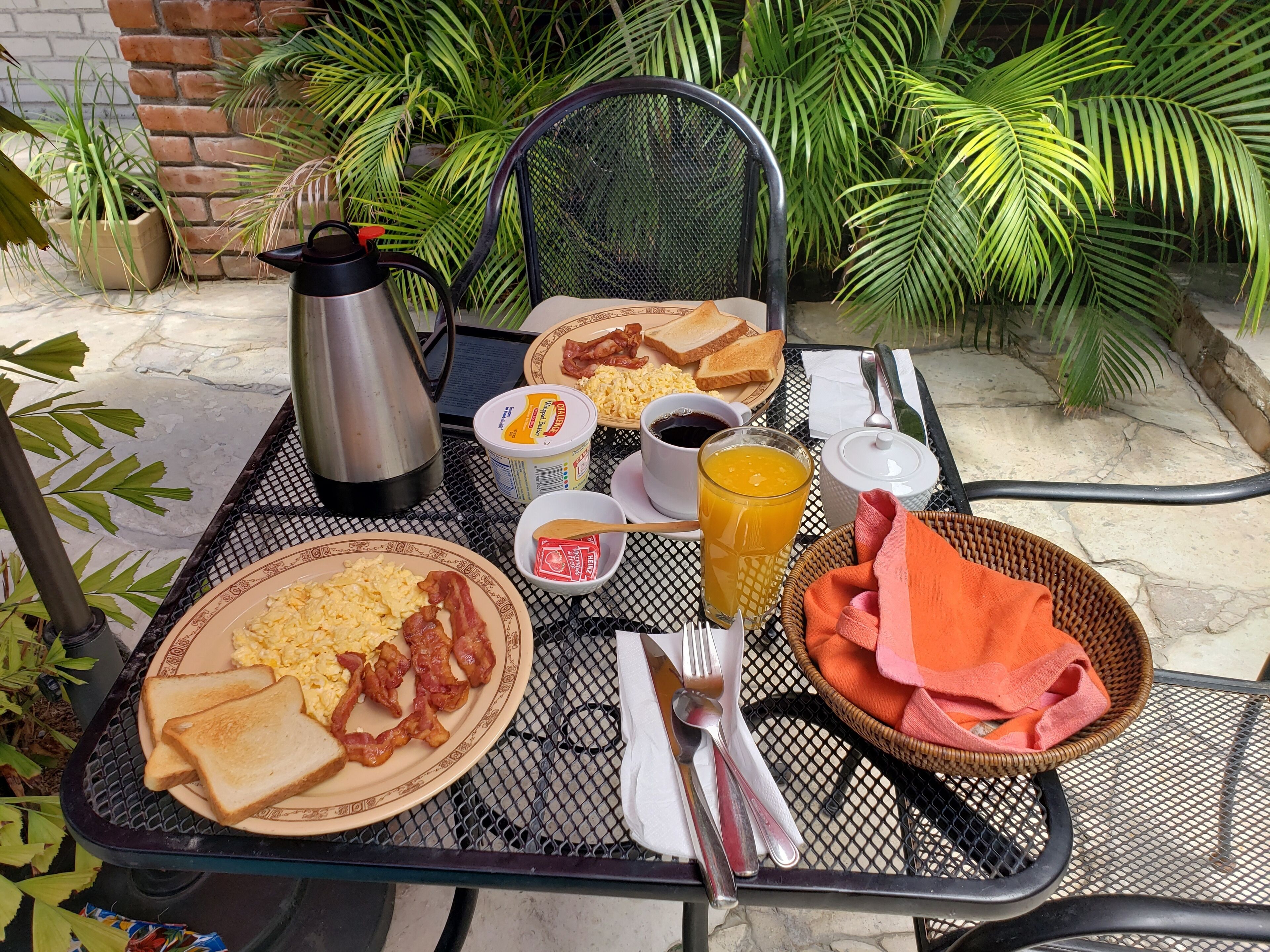 A breakfast table set outdoors with plates of eggs, bacon, toast, and juice, along with a coffee pot and utensils.