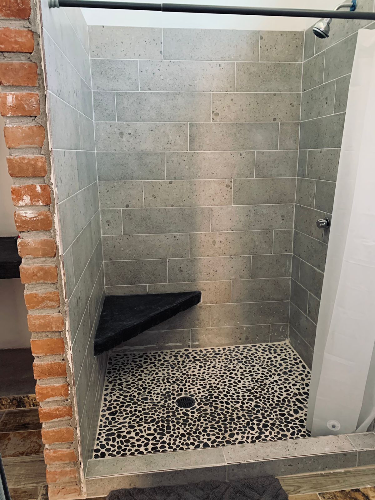 Área de chuveiro com azulejos cinza salpicados, prateleira preta e piso com padrão de pedrinhas.