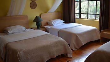 Quarto com duas camas, uma mesa de cabeceira com abajur, uma janela com cortinas e um painel de parede tecido.