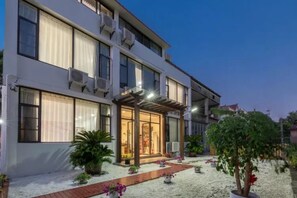 A modern hotel with a covered entrance, large windows, and a well-maintained garden with potted plants and a pathway.