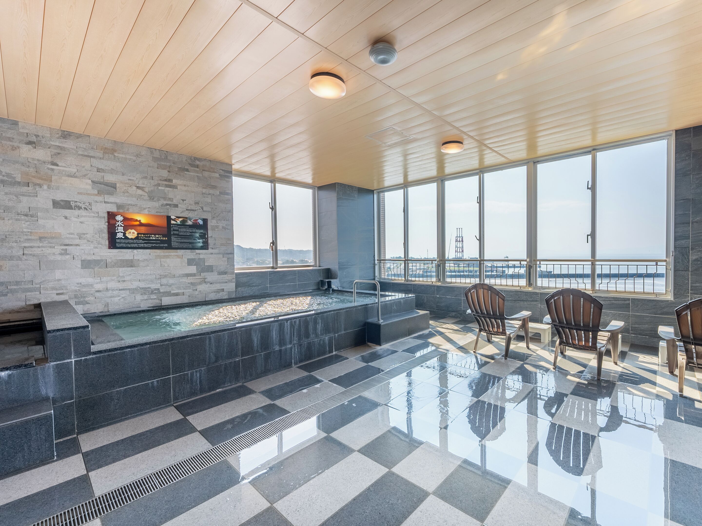 A modern indoor pool area with a stone wall, a hot tub, and a view of the sea through large windows.