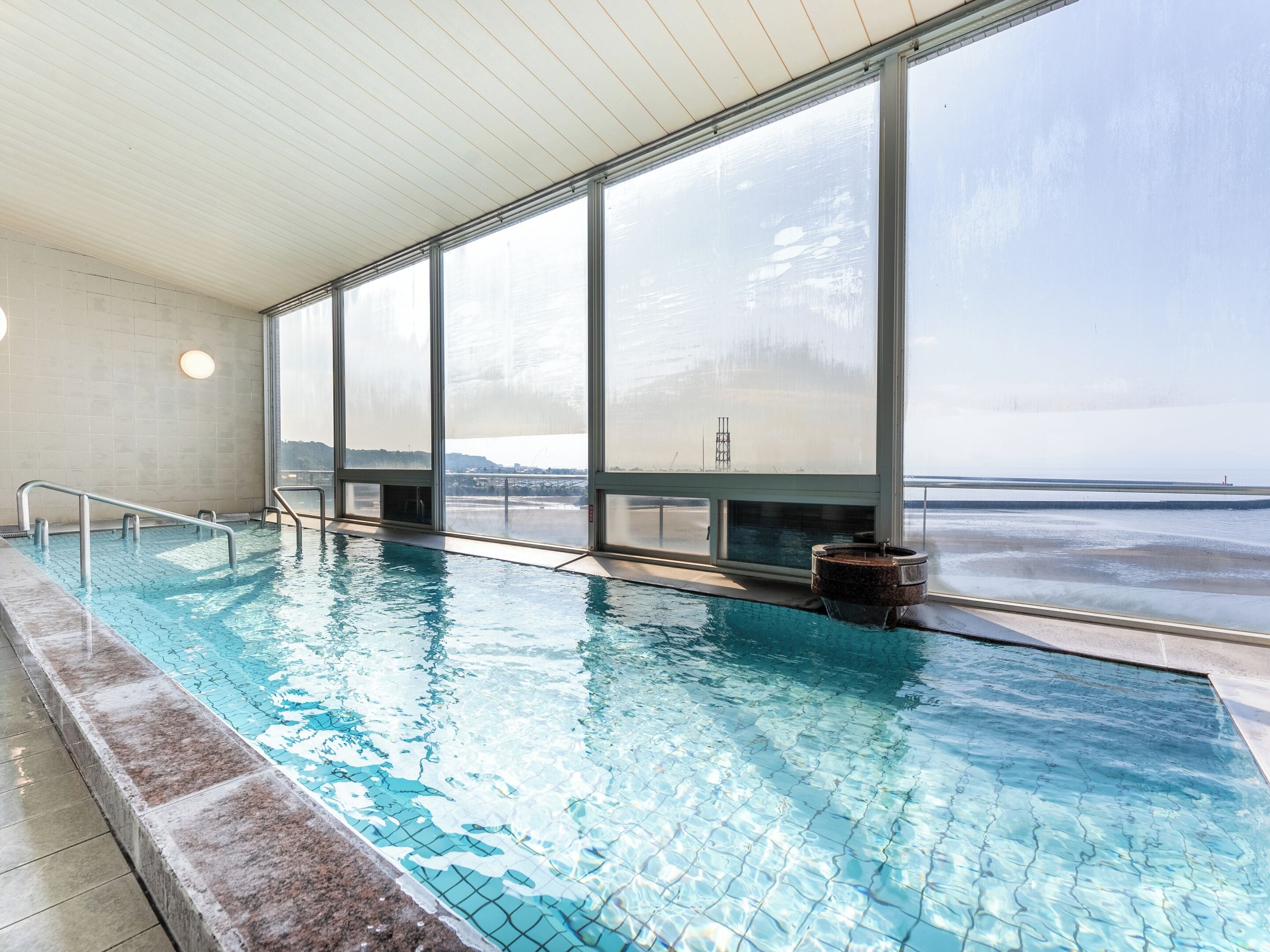 An indoor swimming pool with clear water, large windows, and a view of the beach.