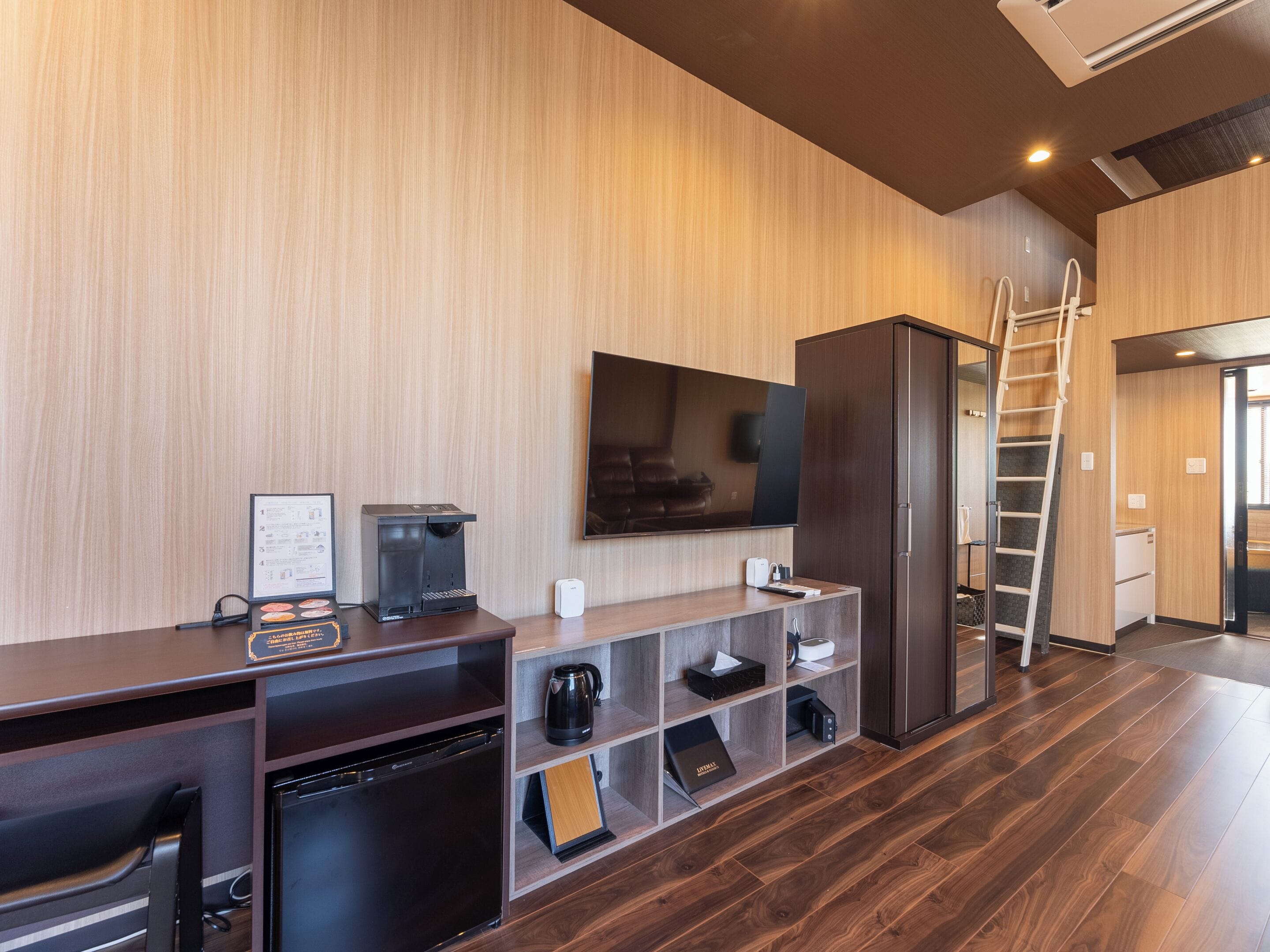 Una habitación de hotel moderna con piso de madera, un escritorio empotrado con cafetera, televisor de pantalla plana y armario.