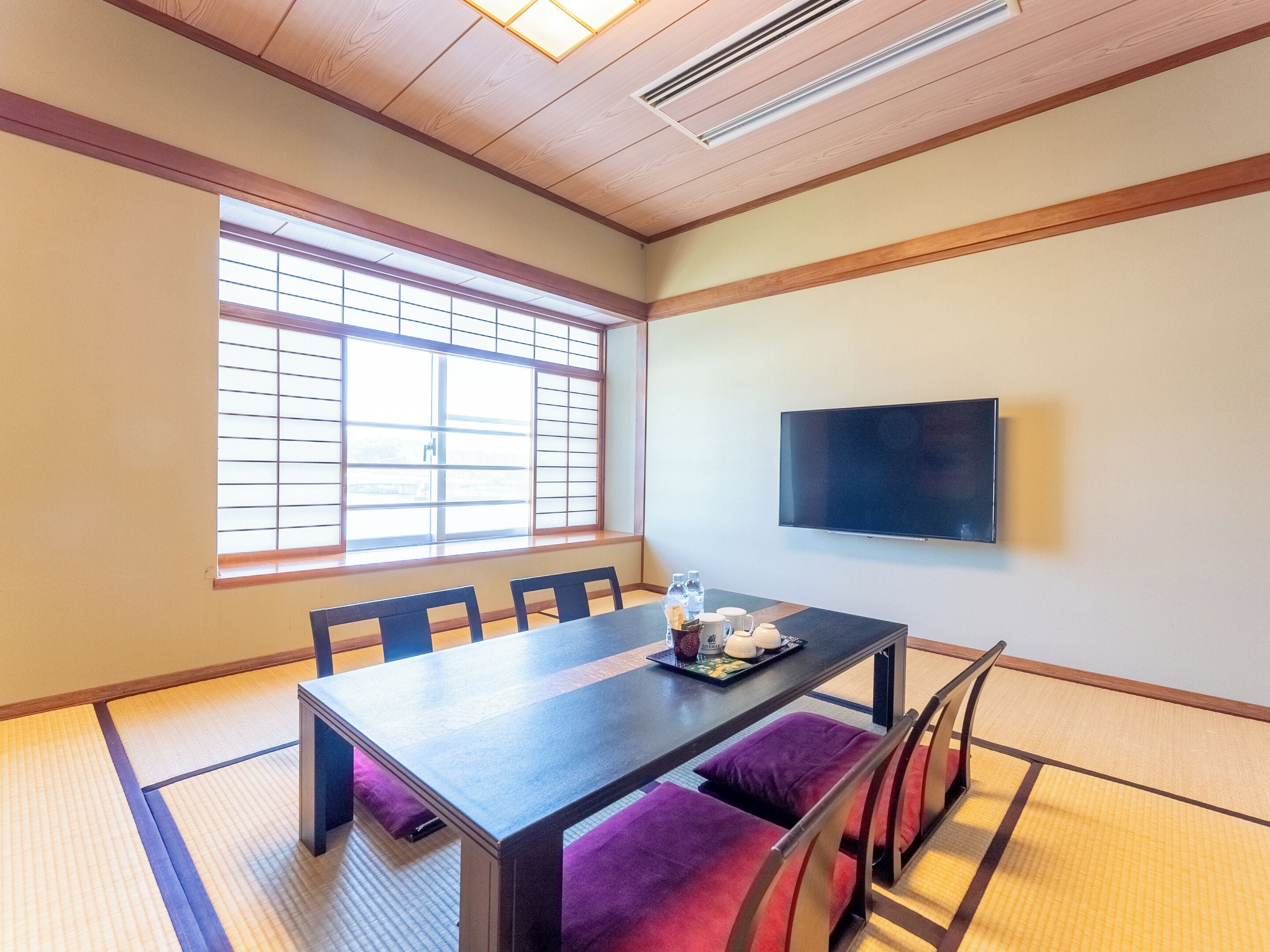 Une pièce de style japonais traditionnel, avec un sol recouvert de tatamis, une petite table basse avec des coussins et un téléviseur fixé au mur.