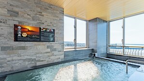 A modern indoor pool with stone walls and large windows offering a view of the city. - LiVEMAX RESORT Sakurajima-seafront (Tarumizu)