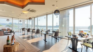 A spacious restaurant with large windows offering a view of a bridge and river. Tables are arranged with chairs, and there are bottles of beer on a wooden stand. - LiVEMAX RESORT Sakurajima-seafront (Tarumizu)