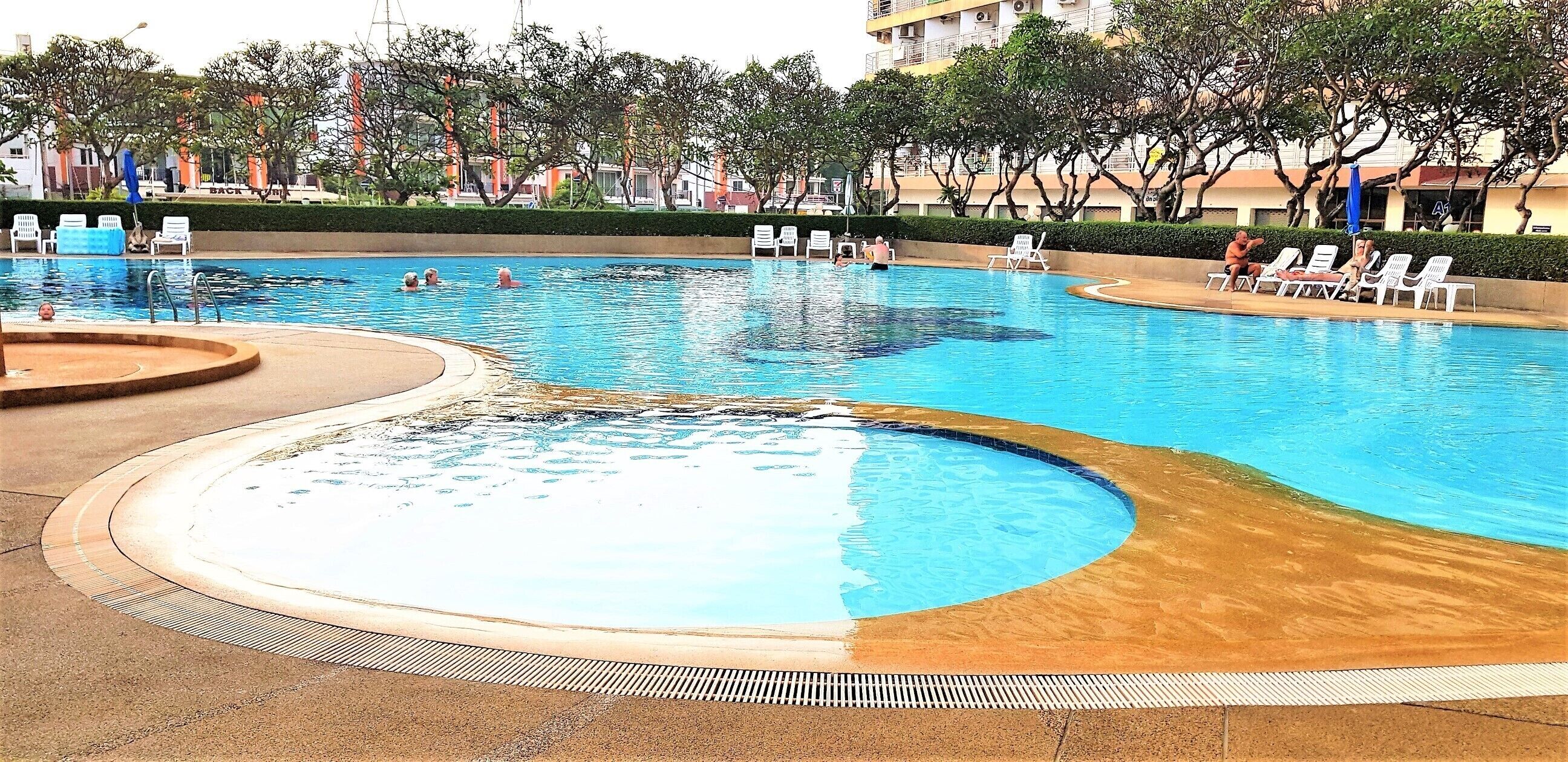 A large outdoor swimming pool with multiple lanes, surrounded by lounge chairs and a landscaped area with trees and a building in the background.
