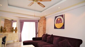 Ein Wohnzimmer mit einem violetten Sofa, einem Holztisch, einem Schreibtisch mit Schubladen und einem Deckenventilator.