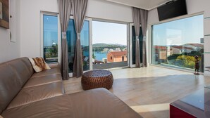 A modern living room with a leather sofa, a round ottoman, and a large glass door leading to a balcony with a view of the sea and buildings. - Villa Martinis (Rogoznica)