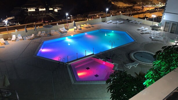 A nighttime view of a swimming pool area with lounge chairs and a smaller pool, surrounded by buildings and streetlights. - Las Brisas de Puerto Rico (Mogán)