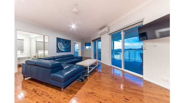 A modern living room with a blue leather sofa, wooden flooring, and a large window offering a view of the sea. - Picturesque on Passage - Whitsundays (Shute Harbour)