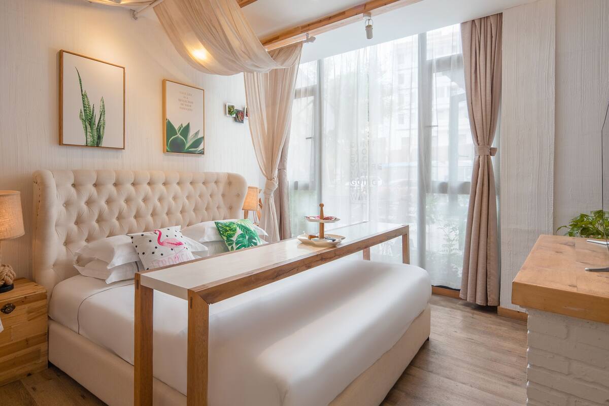 Un dormitorio con cabecera capitonada, una mesita de noche con lámpara, una planta en la mesita y una ventana con cortinas traslúcidas.