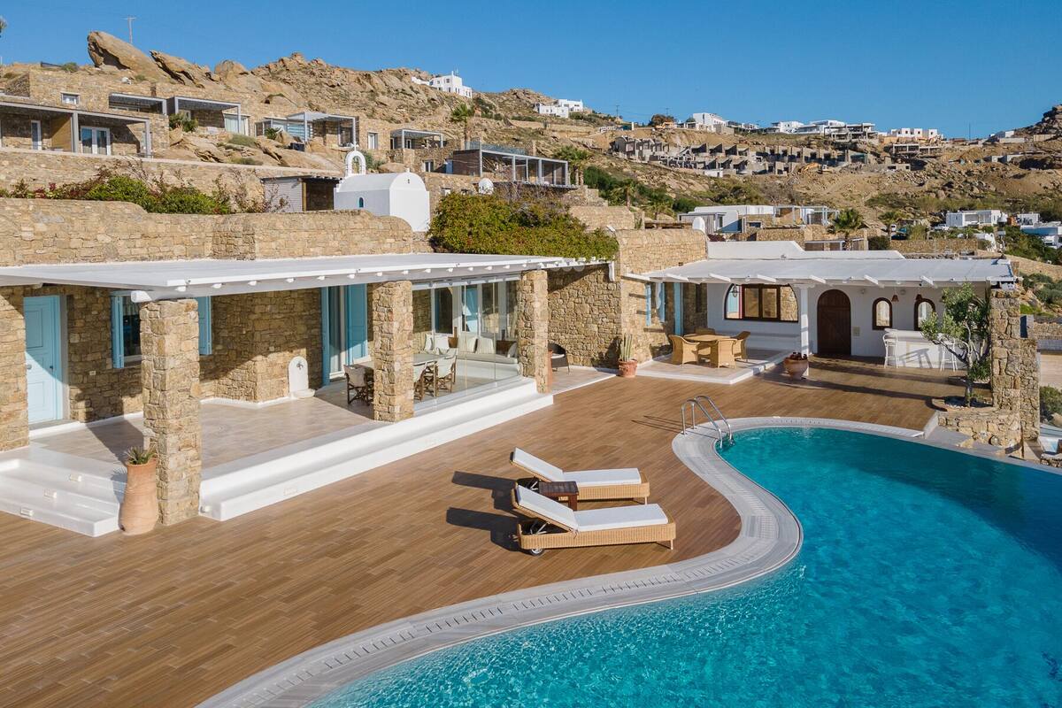 Uma villa moderna com piscina, paredes de pedra e vista para a colina ao redor.