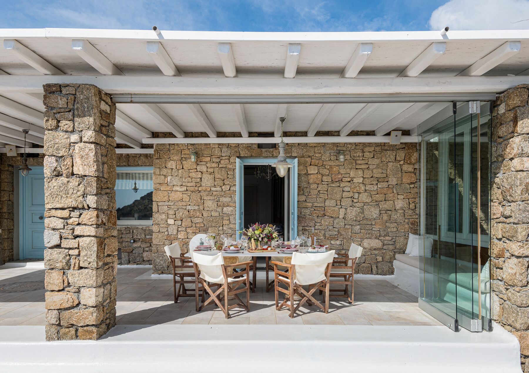 An outdoor dining area with stone walls, a covered pergola, and a glass door leading to the interior.
