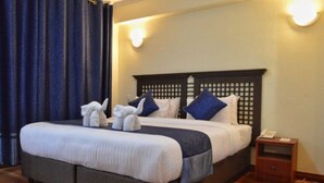 A neatly made bed with a dark headboard, white linens, and blue pillows. There is a small bedside table with a lamp and a phone. - Cranes Suites Hotel (Kitale)