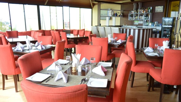 A restaurant with red upholstered chairs and tables, a bar area with shelves of bottles, and large windows providing a view of the outdoors. - Cranes Suites Hotel (Kitale)