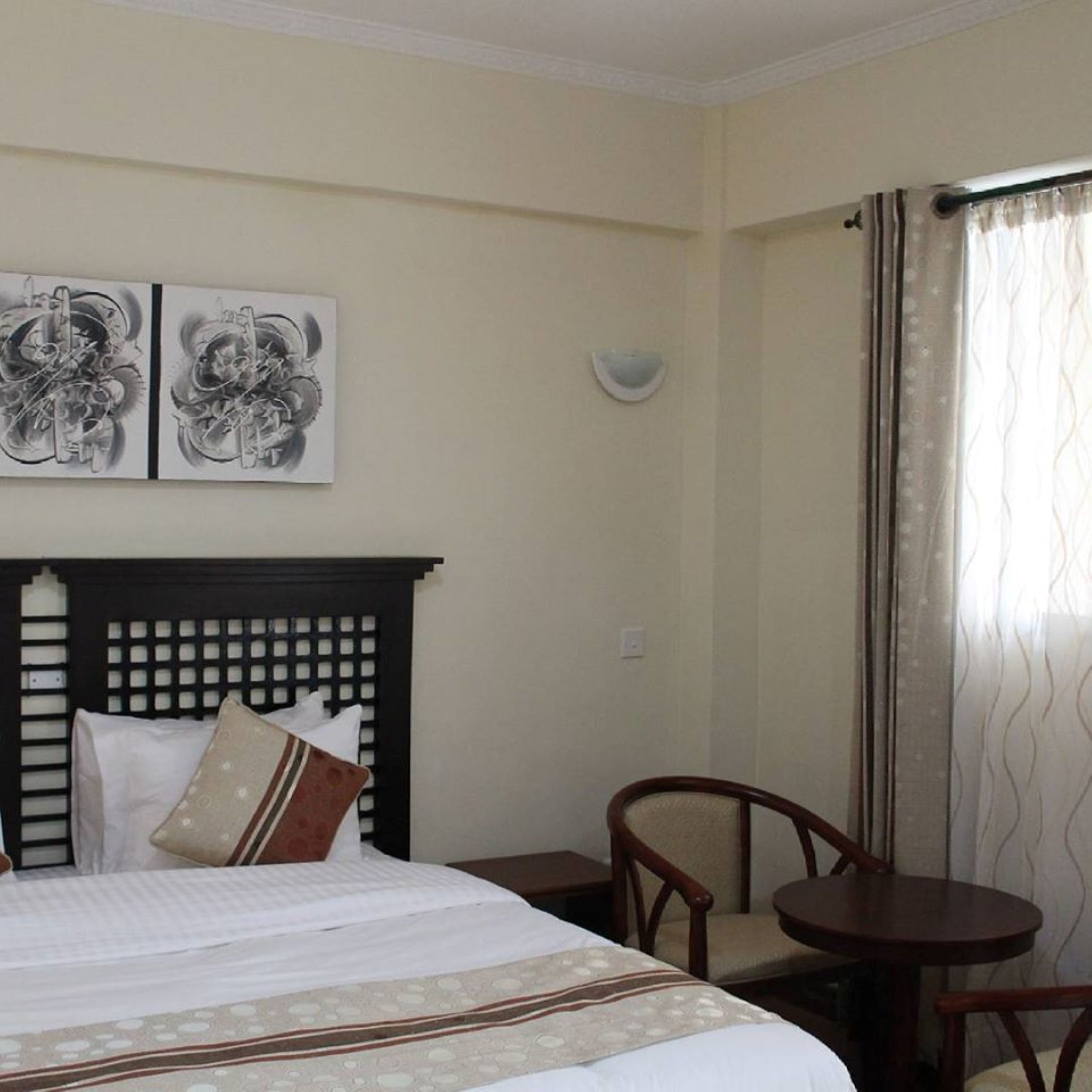 A hotel room with a bed, a headboard, two wall-mounted artworks, a small table, and a chair.