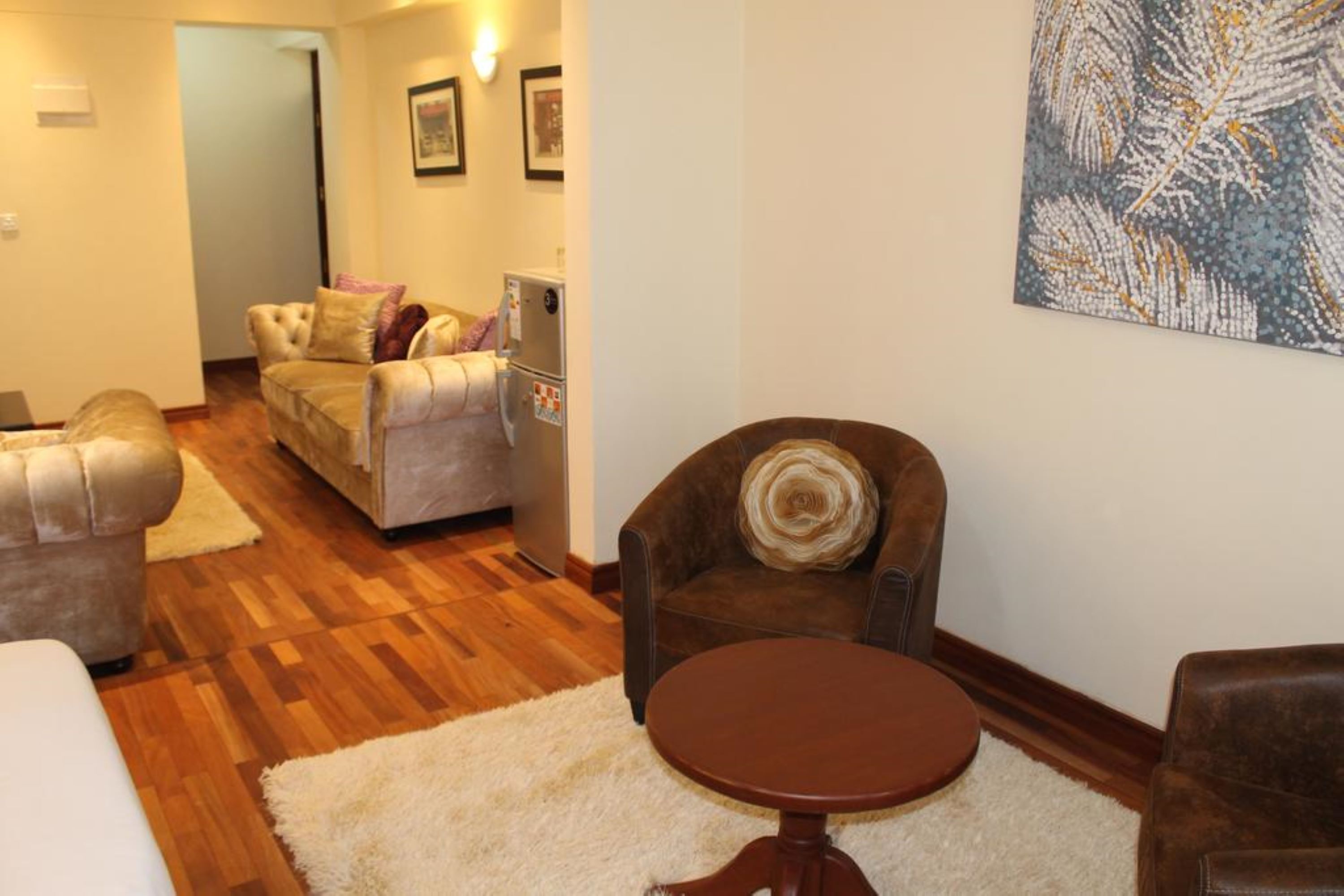 Una sala de estar con un sillón de cuero marrón, un sofá color beige y un tapete blanco. Hay un cuadro en la pared y una puerta que da a otra habitación.