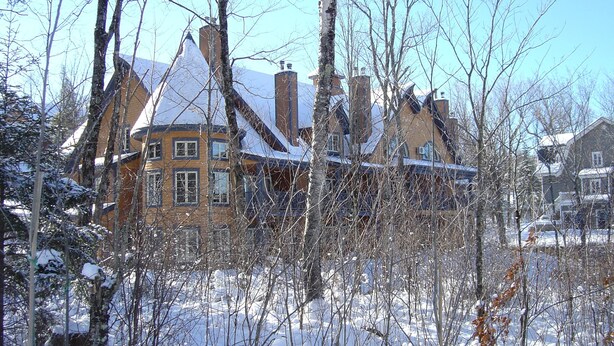A snow-covered house with multiple chimneys, surrounded by leafless trees and a clear sky. - Stunning 2 bed Condo in Les Manoirs, Mont-Tremblant (Mont-Tremblant)