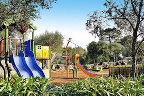 Children can play on the slides and climbing elements in this outdoor playground surrounded by lush greenery and trees.