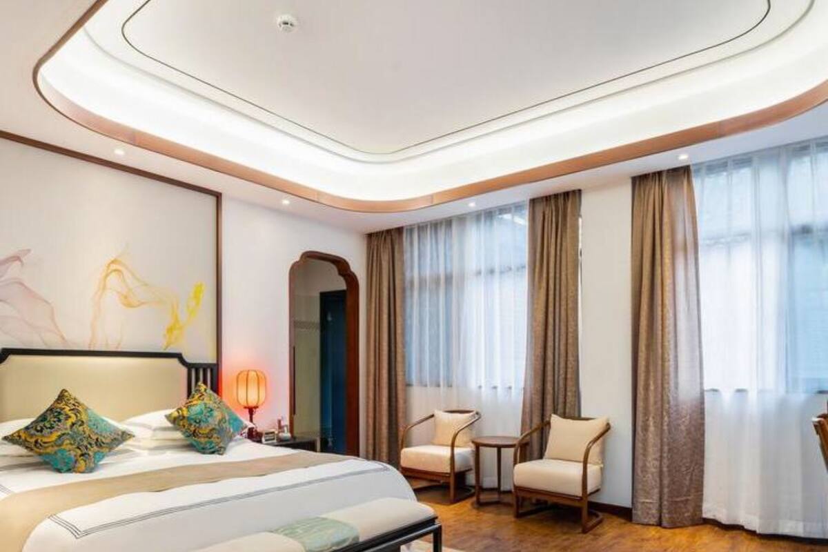 Une chambre d’hôtel moderne avec un grand lit, une banquette, deux chaises, une petite table et une grande fenêtre avec des rideaux.