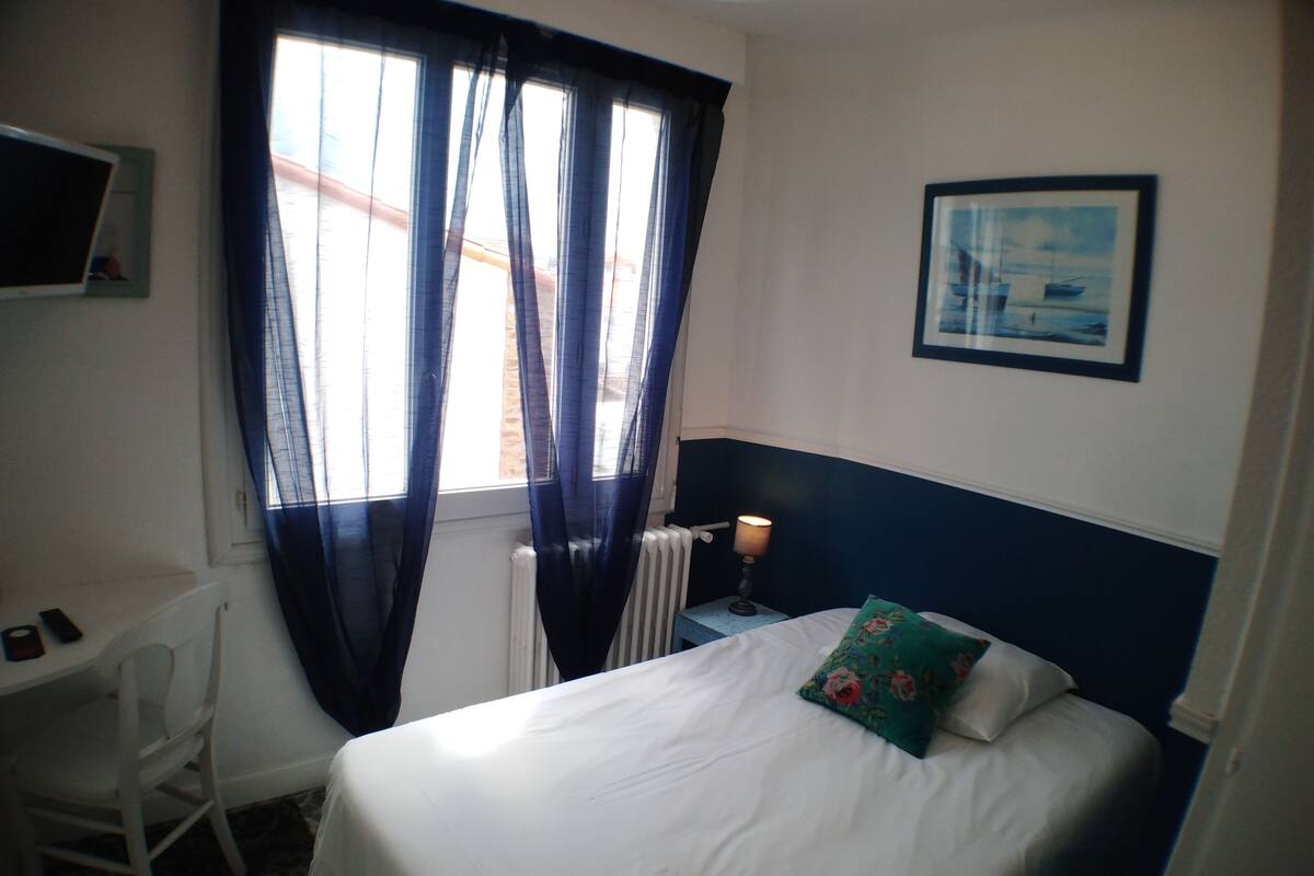 Een hotelkamer met een bed, bureau, stoel, radiator, lamp en een schilderij aan de muur.