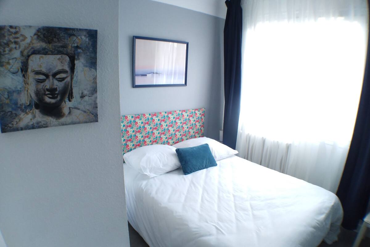 Een net opgemaakt bed met een witte dekbedovertrek en een blauw kussen, een hoofdbord met bloemenmotief, een Boeddha-schilderij aan de muur en een ingelijste foto boven het bed.