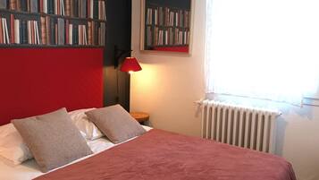 Een slaapkamer met een rode hoofdeinde, een rode lamp, een boekenkast en een raam met gordijnen.