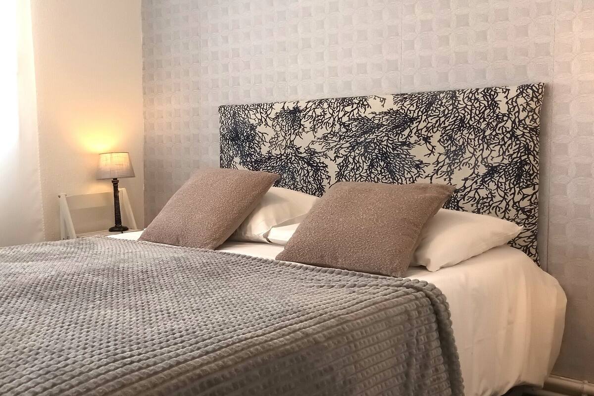 Een opgemaakt bed met een gebreid dekbed, een hoofdbord met patroon en een bedlamp.