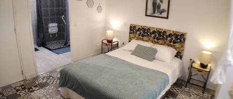 A bedroom with a bed, a nightstand, a wall-mounted lamp, a shower area, and a door leading to another room.