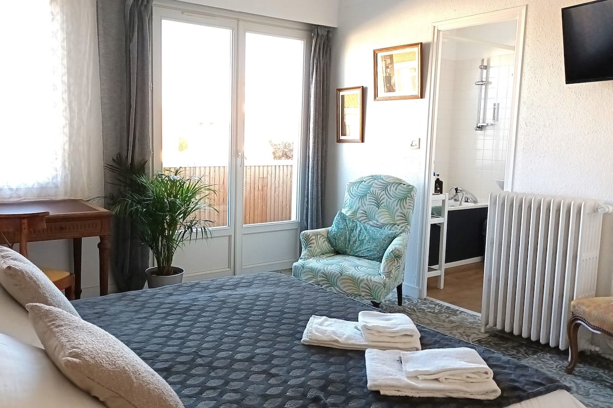 Een slaapkamer met een bed, een stoel, een bureau, een radiator en een raam met gordijnen.