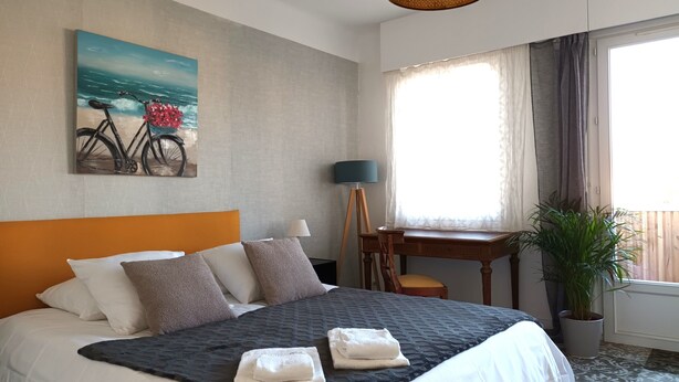 Habitación de hotel con cama, escritorio, silla, lámpara, planta en maceta y un cuadro de bicicleta en la pared.