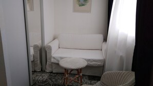 Una habitación pequeña y ordenada con un sofá blanco, una silla de mimbre y un taburete.