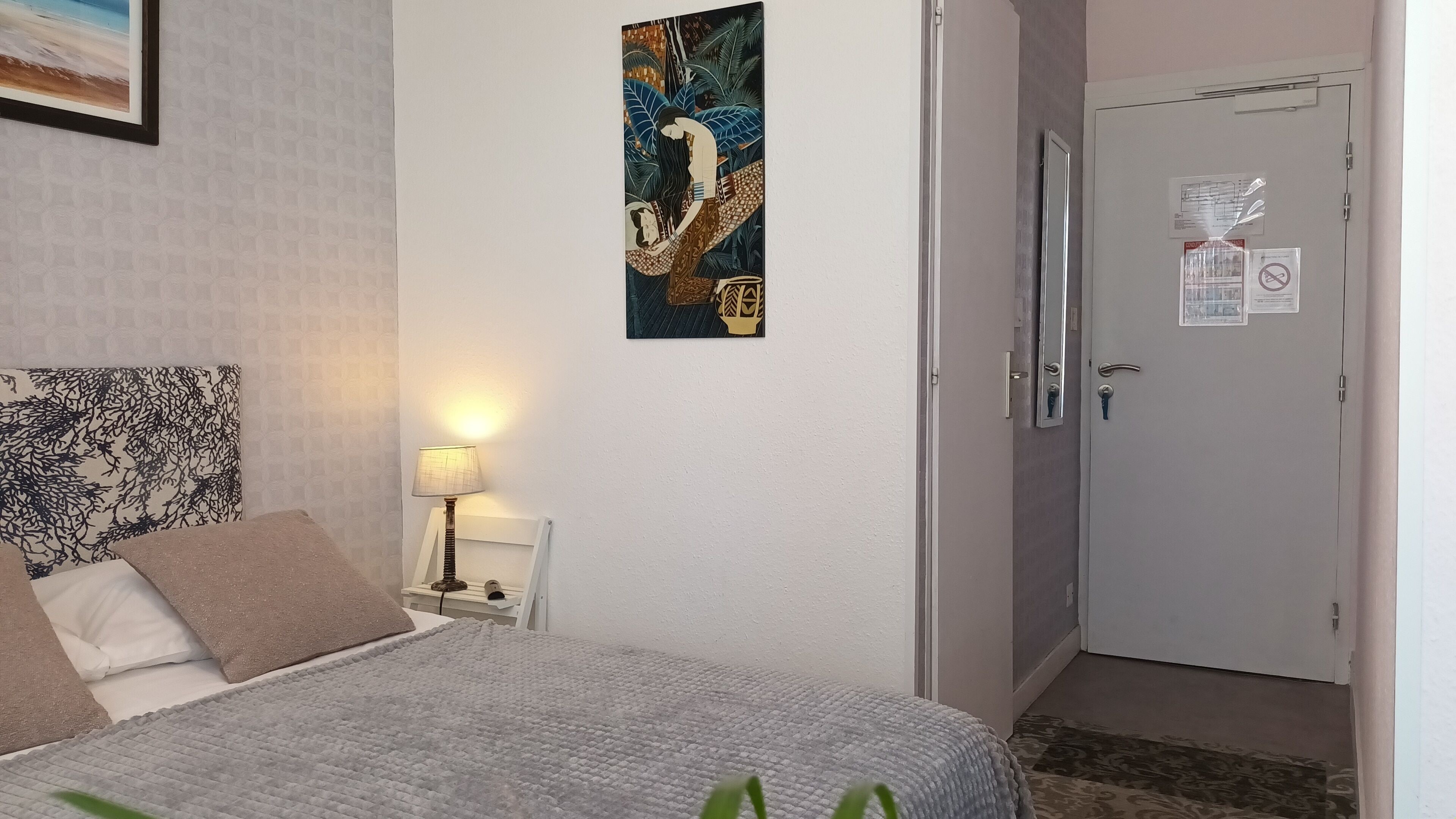 Una habitación de hotel con una cama, una mesita de noche con una lámpara, una obra de arte colgada en la pared, una puerta con espejo y un cartel, y una alfombra estampada.