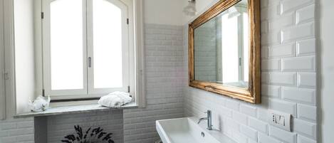 Un baño con paredes de ladrillo blanco, un lavamanos blanco, un espejo con marco de madera y una ventana con una toalla blanca en el alféizar.