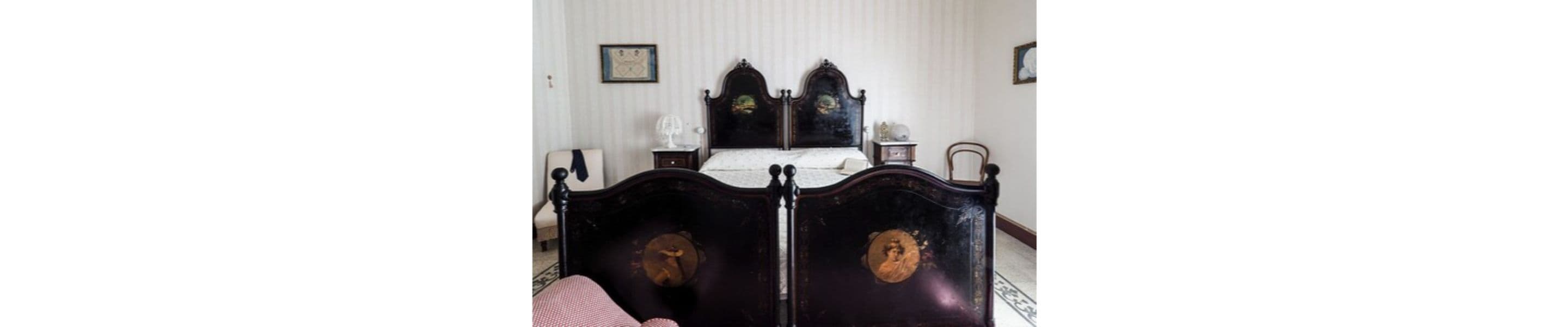 Une chambre à l’ancienne, avec un lit en bois sombre, une chaise et une petite table sur laquelle se trouve une lampe.