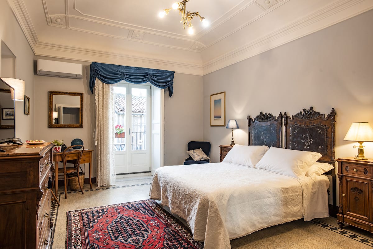 Une chambre à coucher avec un grand lit, une commode en bois, un bureau avec une chaise, une fenêtre avec des rideaux et un lustre.