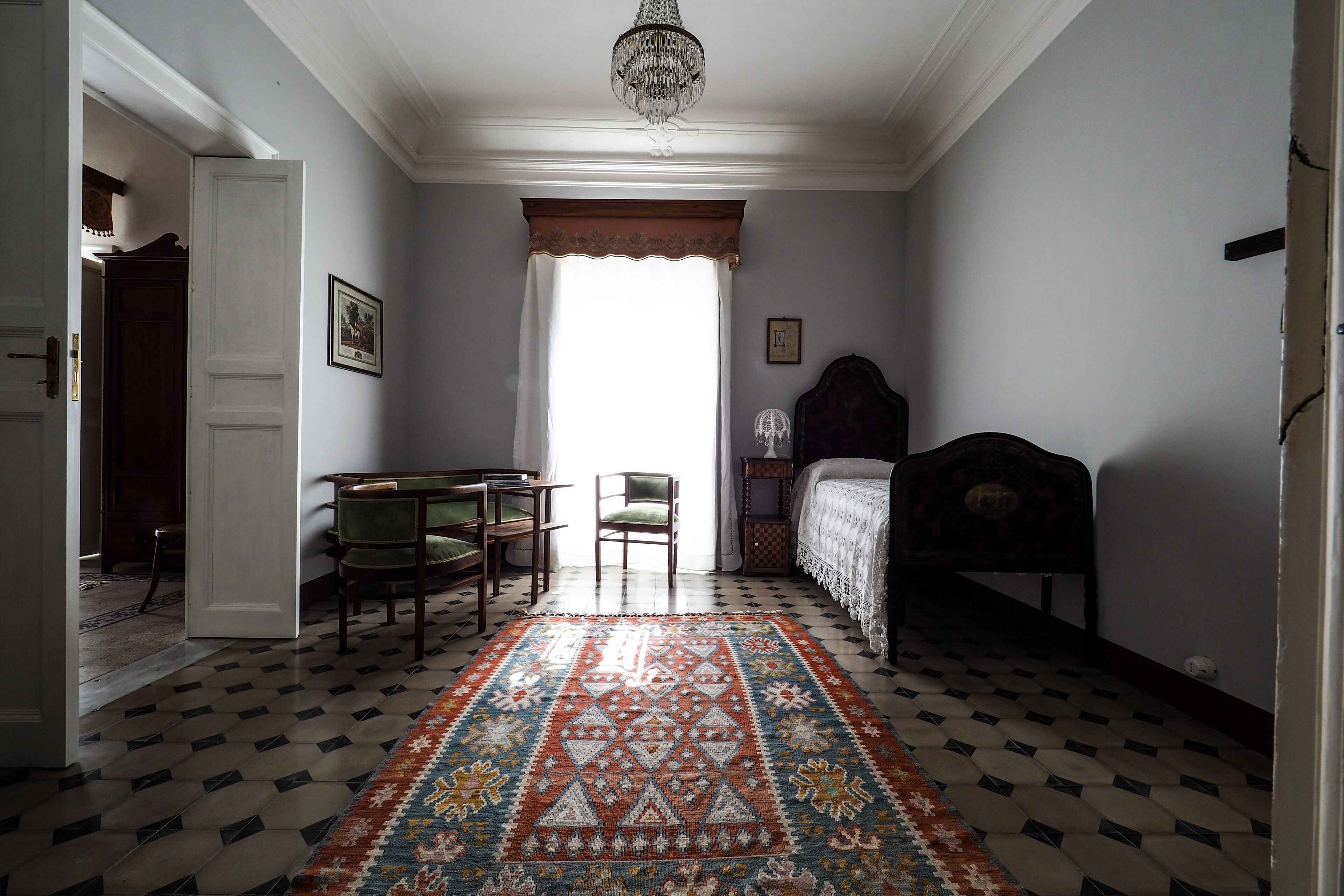 A room with a patterned rug, a bed, a chair, a desk, and a chandelier.