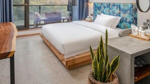Ein modernes Hotelzimmer mit einem großen Bett, einem Schreibtisch mit Spiegel, einer Zimmerpflanze und Blick ins Freie.