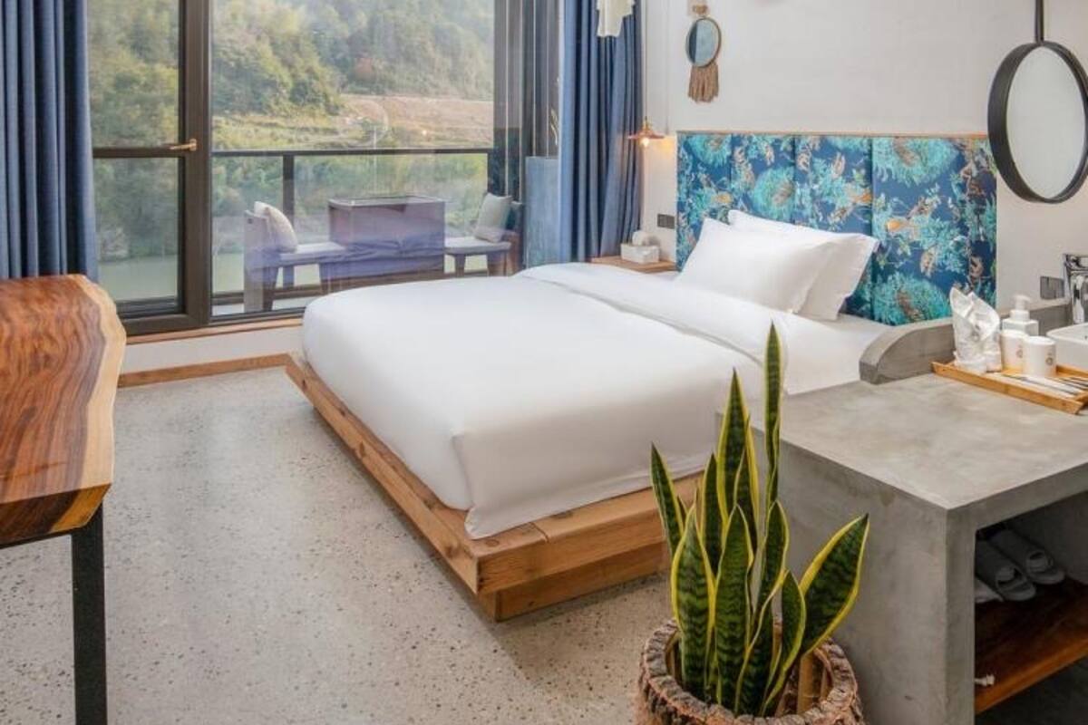 Une chambre d’hôtel moderne avec un grand lit, un bureau doté d’un miroir, une plante en pot et une vue sur l’extérieur.