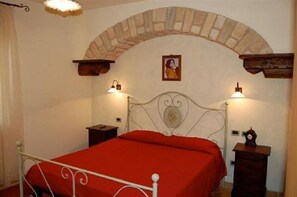 Ein Schlafzimmer mit roter Bettdecke, einem weißen Metallbettgestell, einem Steinbogen, zwei Nachttischen mit Lampen und einem gerahmten Bild an der Wand.